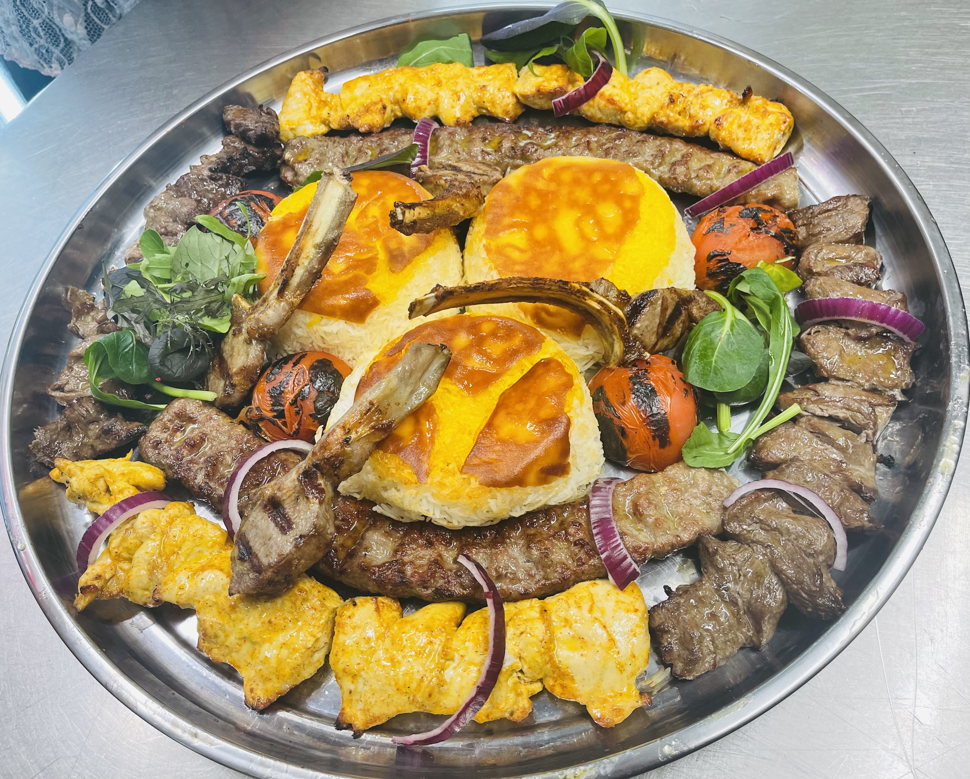 A huge round silver platter packed with a full mixed grill — koobideh, saffron joojeh chicken, sliced barg, charred tomatoes and herb salad