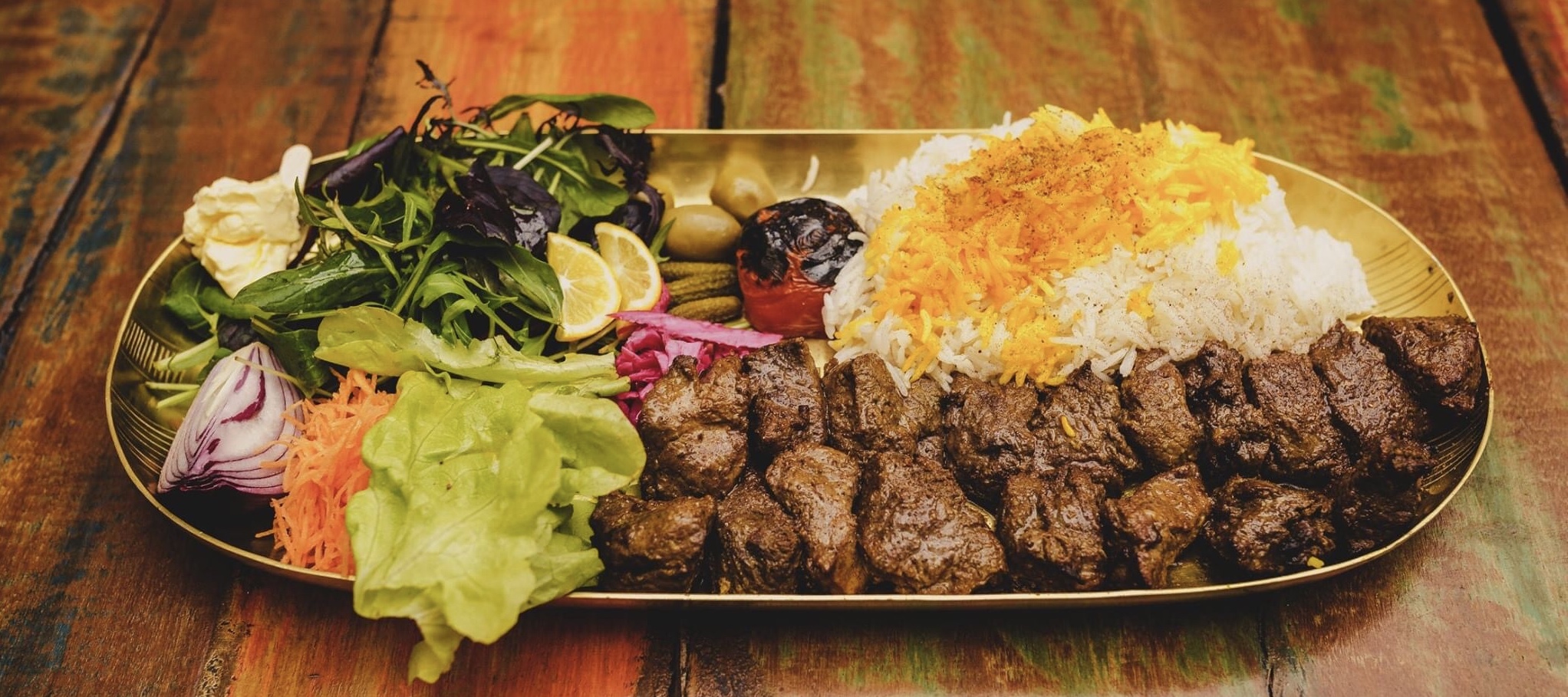 A long gold platter of grilled chenjeh chunks beside saffron rice with a fan of fresh salad and herbs